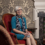 Photograph of Alison Rose, sitting in a red armchair, wearing a blue dress and patterned scarf, and smiling at the camera