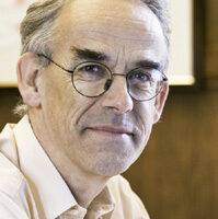 Colour head and shoulders photo of Sir Christopher Hum, wearing glasses and a light shirt