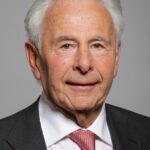 Colour head and shoulders portrait photograph of Lord Levy, wearing a grey suit and red and silver tie