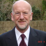 Head and shoulders photo of Derek Marsh, wearing glasses, navy suit and maroon tie, with garden wall in background